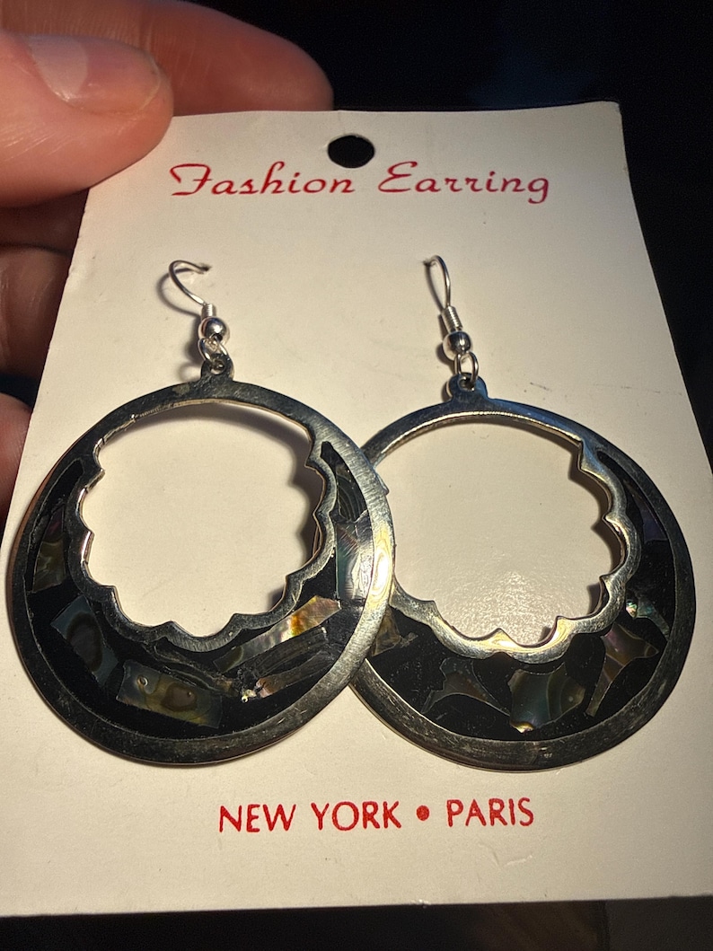 Op de afbeelding: Zilverkleurige oorringen met een geschulpte rand en ingelegde zwarte en iriserende schelpstukken. De oorbellen worden getoond op een witte kaart met de tekst "Fashion Earring" en "NEW YORK &bull; PARIS".