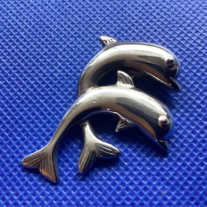 May include: A silver-toned dolphin brooch featuring two dolphins in a playful pose. The brooch has a polished finish, with small red accents for the eyes, set against a textured blue background.