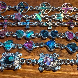 May include: A collection of six silver-toned link bracelets featuring inlaid iridescent abalone shell accents. The designs include peace signs, stars, crosses, butterflies, hearts, and turtles, each connected by small metal links in a row.