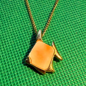May include: A gold-toned necklace featuring a pendant shaped like a fish with a smooth, light orange, square-like centre inlay. The pendant is suspended from a delicate gold-toned chain, showcasing a metallic finish and a distinct fish silhouette.
