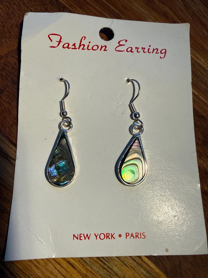 Op de afbeelding: Een paar druppelvormige oorbellen met iriserende abalone schelpinzetstukken, gezet in zilverkleurig metaal. De oorbellen worden gepresenteerd op een witte kaart met de tekst "Fashion Earring" en "NEW YORK &bull; PARIS".