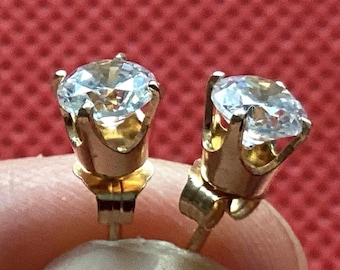 May include: A pair of gold stud earrings, each featuring a single round, clear, faceted gemstone held in a four-prong setting. The earrings have a polished gold finish and include standard post backs for secure attachment to the earlobe.