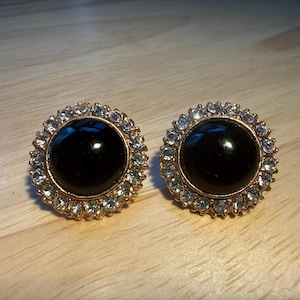 May include: A pair of gold-toned stud earrings. Each earring features a large, round, black center stone surrounded by a halo of small, clear crystals. The earrings are set against a light-colored wooden surface.