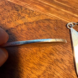 May include: A silver pendant with a circular design is shown in close-up. The pendant's edge is visible, held by a hand, with a wooden surface in the background. The pendant has a shiny, reflective surface.