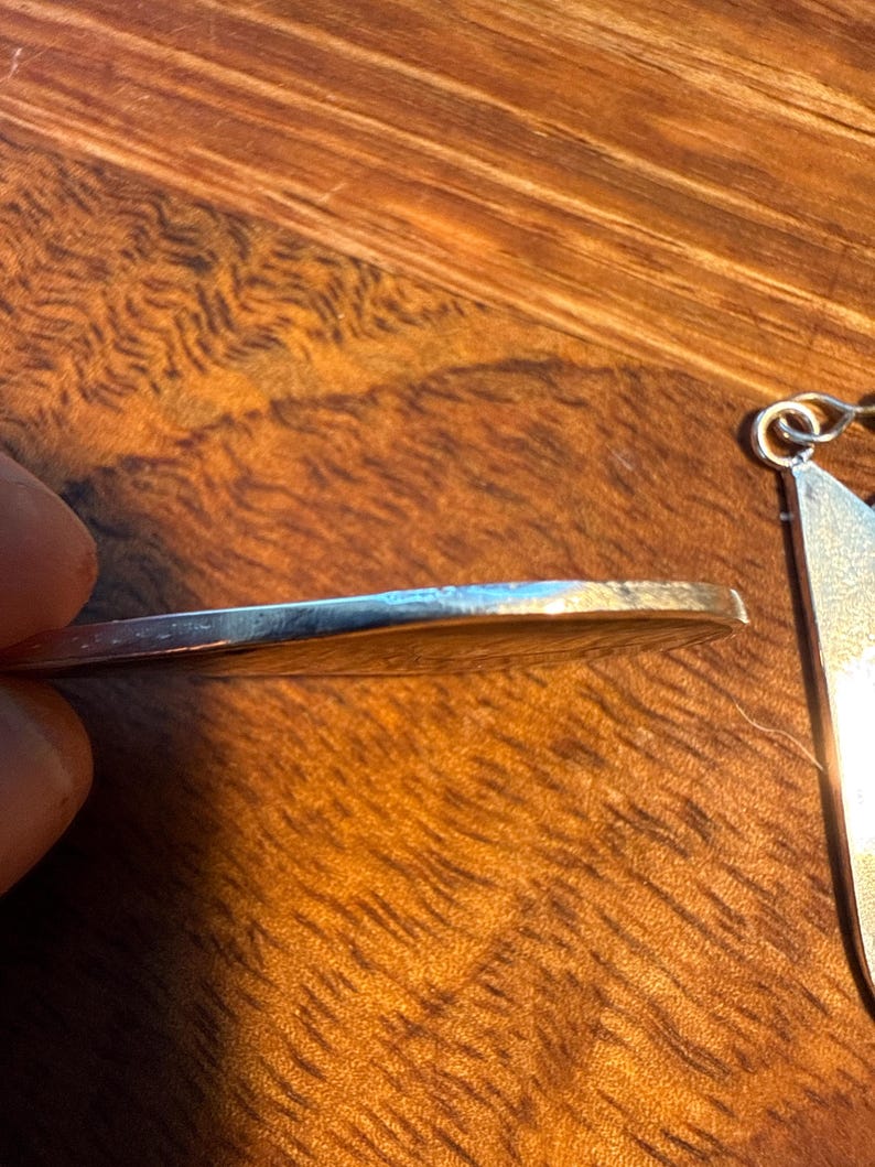 May include: A silver pendant with a circular design is shown in close-up. The pendant's edge is visible, held by a hand, with a wooden surface in the background. The pendant has a shiny, reflective surface.