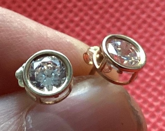 May include: A pair of gold-tone stud earrings, each featuring a single round, clear, faceted stone set in a circular bezel setting with an open gallery design. The earrings are held in place, showcasing the reflective quality of the stones.