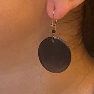 May include: A dark brown, circular earring with a gold-colored hook and small black beads. The earring is a simple, minimalist design, with a smooth, polished surface. The earring is shown on a person's ear.