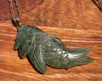 Puede incluir: Un colgante de piedra verde oscuro tallado con forma de pez con escamas y aletas detalladas, suspendido de una cadena de cuerda trenzada en tono dorado. El colgante presenta una superficie lisa y pulida y está unido a la cadena por una argolla.