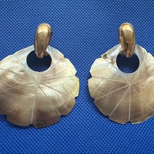 May include: A pair of earrings featuring a gold-coloured top and a fan-shaped, mother-of-pearl design. The earrings have a circular hole in the centre and are set against a blue background. The earrings are approximately 5 cm in diameter.