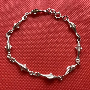 Puede incluir: Una pulsera de plata con dijes de delfines. La pulsera está hecha de segmentos unidos, cada uno con un diseño de delfín. La pulsera se muestra sobre un fondo rojo.