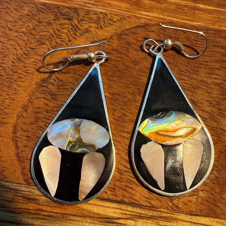 May include: A pair of teardrop earrings featuring black enamel and iridescent abalone shell inlays. The earrings have a silver-tone metal frame and ear wires. The bottom of the earrings have two light pink shell inlays.