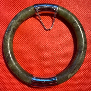 Könnte beinhalten: Ein runder Jade-Armreif mit einer gesprenkelten grünen Farbe. Das Armband hat zwei silberfarbene Metallverschlüsse, die durch eine kleine Kette verbunden sind. Das Armband wird auf einer roten Oberfläche präsentiert.