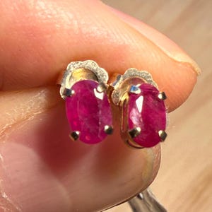 Puede incluir: Un par de pendientes de oro con forma de botón, cada uno con una gema de rubí ovalada. Los rubíes muestran un rico tono rojo intenso, engastados en un engaste clásico de cuatro puntas. Los pendientes tienen un delicado respaldo de oro festoneado.