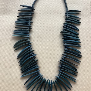 Puede incluir: Un collar de color turquesa con una serie de colgantes curvos en forma de hoja. Los colgantes están ensartados con un hilo de cuentas, creando un diseño único. El collar tiene un cierre magnético.