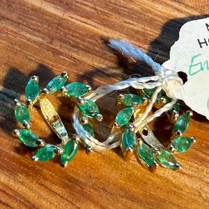 May include: A pair of gold earrings featuring a circular wreath design set with marquise-cut green gemstones. The earrings are connected by a white string and include a small white paper tag with handwritten text partially visible in the frame.