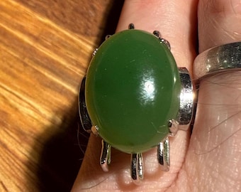 May include: A silver ring featuring a large, oval-shaped, polished green stone. The stone is set in a silver band with decorative prongs. The ring is shown on a finger, with another silver ring visible in the background.