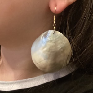 May include: A pair of large, round, white shell earrings with a gold hook. The earrings are hanging from the earlobe.