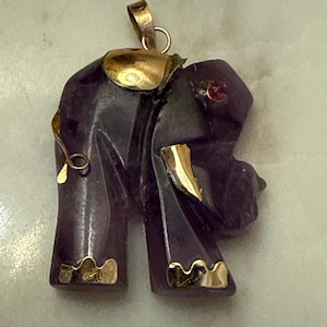May include: A purple elephant pendant with gold accents. The pendant features a carved elephant design with gold details on the ears, feet, and trunk. A small red gem is set in the elephant's eye. The pendant has a gold bail.