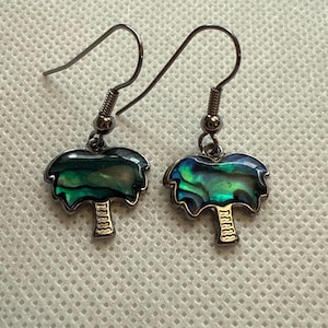 Peut inclure: Une paire de boucles d'oreilles avec un motif de palmier. Les boucles d'oreilles ont une finition en métal argenté avec une fermeture à crochet. Le motif de palmier a une incrustation colorée en coquille d'ormeau, avec des nuances de vert, de bleu et de violet.