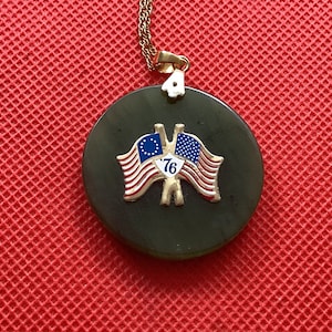 Può includere: Una collana con un ciondolo in giada verde e una catena d'oro. Il ciondolo presenta due bandiere incrociate, una con il design di Betsy Ross e l'altra con la bandiera americana moderna. Le bandiere sono circondate da un '76' dorato su uno sfondo verde.