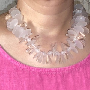 Puede incluir: Un collar blanco opaco de plástico hecho de muchas pequeñas cuentas planas de forma ovalada. Las cuentas están dispuestas en un patrón en capas en cascada.