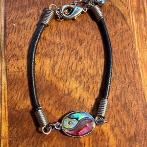 May include: A black leather bracelet with a silver clasp and an oval yin-yang centrepiece. The centrepiece has iridescent, red, and yellow accents. Silver wire wraps the leather. The bracelet is a stylish accessory.