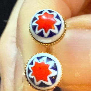 May include: A pair of round stud earrings with a gold-colored border. Each earring features a white background with a red starburst design in the center, outlined by blue and white shapes. The earrings are small and delicate.