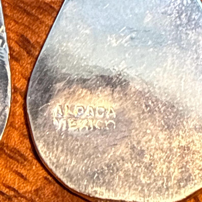 May include: Close-up of a silver earring with a teardrop shape. The text "ALPACA MEXICO" is stamped on the surface. The earring is set against a warm, brown background.