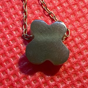 May include: A gold-colored chain necklace with a four-leaf clover pendant. The pendant is a muted green color and has a smooth, rounded shape. The necklace is displayed against a red textured background.