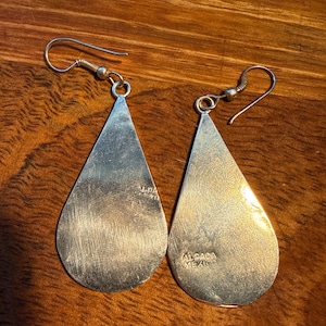 May include: A pair of silver teardrop earrings with a hook fastening. The earrings have a polished, reflective surface and are stamped with "ALPACA MEXICO". The earrings are displayed on a wooden surface.