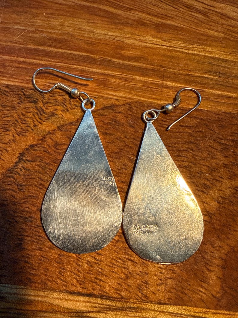 May include: A pair of silver teardrop earrings with a hook fastening. The earrings have a polished, reflective surface and are stamped with "ALPACA MEXICO". The earrings are displayed on a wooden surface.