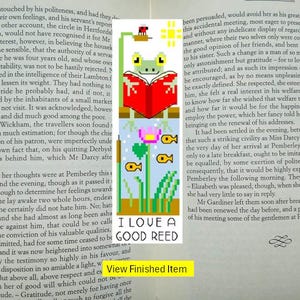 以下が含まれることがあります： 「I Love a Good Reed」の文字が入った、本を読むカエルのピクセルアートのブックマーク。デザインには花と魚が含まれています。読書好きに最適です。