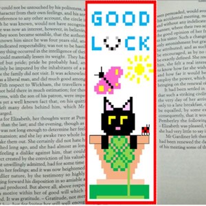 Puede incluir: Un marcapáginas pixelado con las palabras "GOOD LUCK" en azul. Presenta una mariposa rosa, un sol amarillo y un gato negro en una maceta. Una mariquita roja está a la derecha del gato.