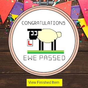 May include: A circular embroidery hoop containing white fabric with a cross-stitched design of a sheep standing on a green patch. The sheep holds a white square with a red letter L. The text CONGRATULATIONS and EWE PASSED is stitched in black.