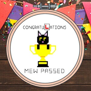 May include: A circular wooden embroidery hoop containing white fabric with a cross-stitch design. The design features a black pixelated cat inside a yellow trophy cup, with the text CONGRATULATIONS and MEW PASSED in black lettering.