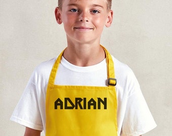 Può includere: Un grembiule giallo brillante con il nome "ADRIAN" stampato in lettere nere in grassetto. Il grembiule ha un cinturino per il collo e una fibbia. La persona indossa una maglietta bianca e jeans blu.