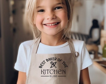 Può includere: Un grembiule beige con la scritta "BEST BAKER OF THE KITCHEN" e il nome "ELISE" in nero. Il grembiule presenta un disegno di frusta e mattarello. Il grembiule è indossato da un bambino sorridente con una maglietta bianca.