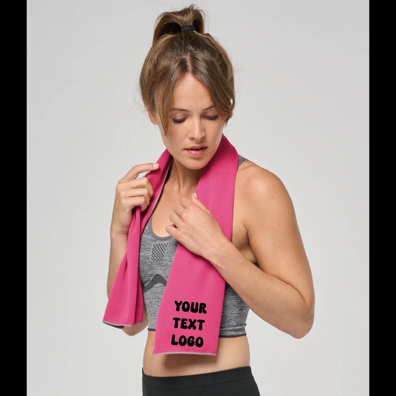 Op de afbeelding: Een roze sportdoek met de zwarte tekst "YOUR TEXT LOGO". De doek is om de nek van een persoon gedrapeerd die een grijze sportbeha en een zwarte sportbroek draagt. De persoon heeft haar haar in een paardenstaart.