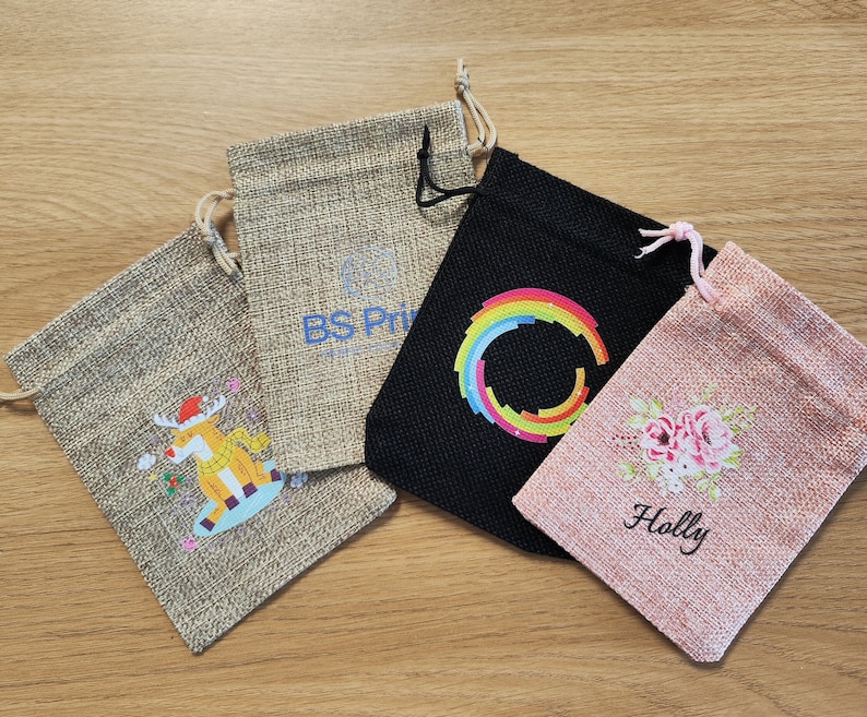 Puede incluir: Cuatro peque&ntilde;as bolsas de arpillera con diferentes dise&ntilde;os. Una bolsa tiene un reno con un gorro rojo, otra tiene un c&iacute;rculo arco&iacute;ris, otra tiene un dise&ntilde;o floral con la palabra "Holly" y la &uacute;ltima bolsa tiene el texto "BS Prints".