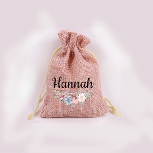 Puede incluir: Una bolsa de yute rosa con cordón de ajuste con el nombre "Hannah" impreso en letra negra. Debajo del nombre hay un diseño floral con flores rosas, azules y verdes.