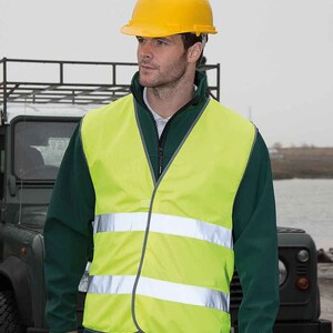 Op de afbeelding: Een gele veiligheidsvest met reflecterende strepen. De vest wordt gedragen over een groene jas en een gele helm.