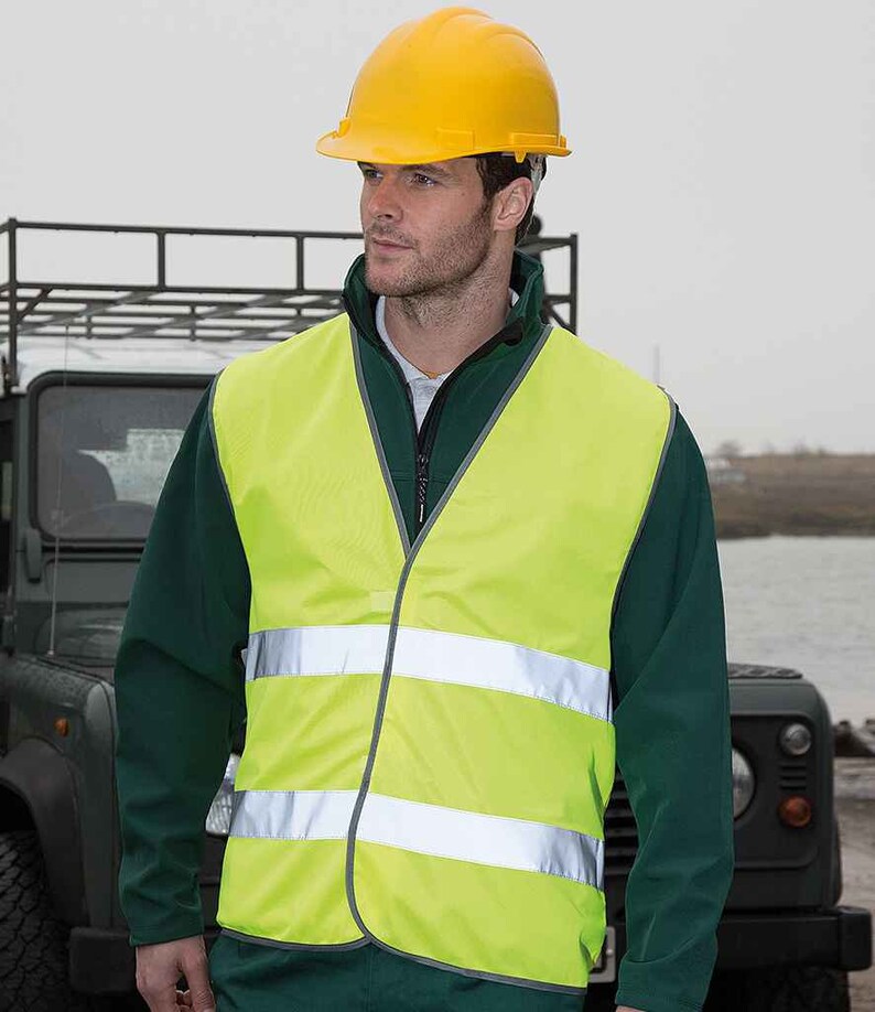 Op de afbeelding: Een gele veiligheidsvest met reflecterende strepen. De vest wordt gedragen over een groene jas en een gele helm.