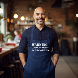 May include: A navy blue apron with the text "WARNING! DAD COOKING IN PROGRESS" in white. The apron is worn by a smiling man in a dark blue shirt. The background is a restaurant setting with warm lighting.