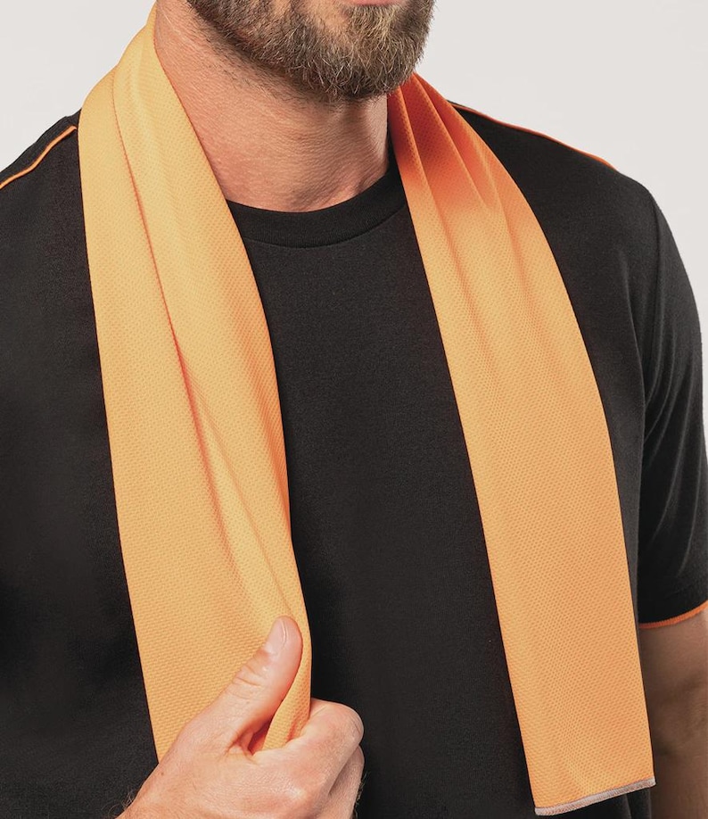 Op de afbeelding: Een oranje koelhanddoek gedrapeerd om de nek van een persoon. De handdoek heeft een gestructureerd oppervlak en wordt gecombineerd met een zwart t-shirt met oranje afwerking. De handdoek is gemaakt van lichtgewicht, ademend materiaal.
