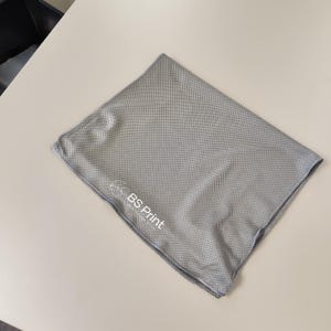 Op de afbeelding: Een grijze, vierkante microvezel handdoek met een geperforeerde textuur. De handdoek heeft een logo met de tekst "BS Print" in het wit. De handdoek is opgevouwen en ligt op een lichtgekleurd oppervlak.