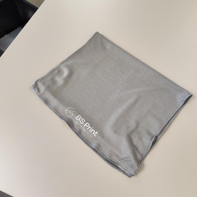 Op de afbeelding: Een grijze, vierkante microvezel handdoek met een geperforeerde textuur. De handdoek heeft een logo met de tekst "BS Print" in het wit. De handdoek is opgevouwen en ligt op een lichtgekleurd oppervlak.