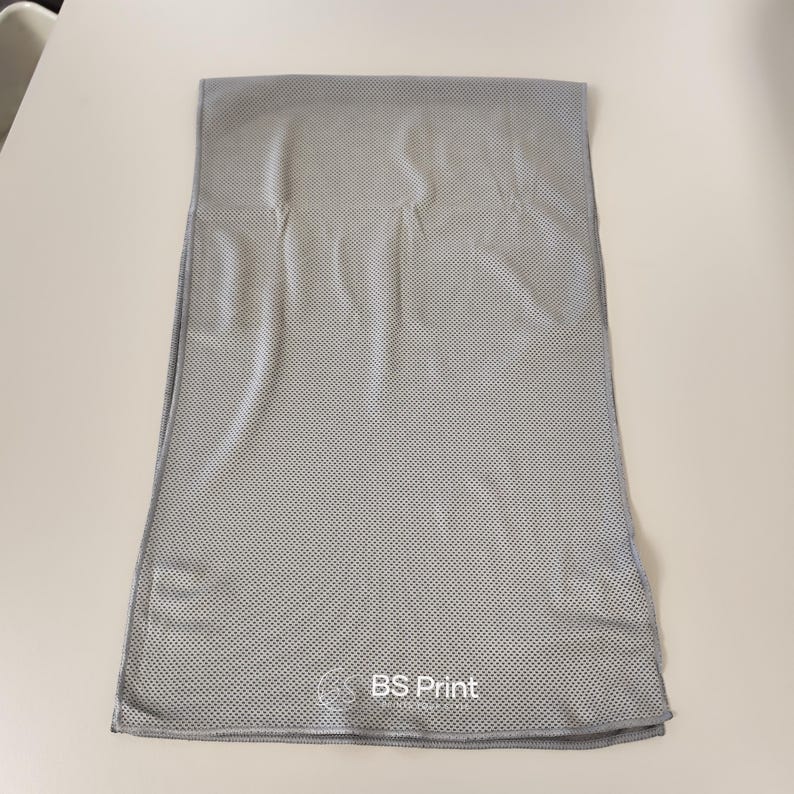 Op de afbeelding: Een grijze, rechthoekige koelhanddoek met een geperforeerde textuur. De handdoek is opgevouwen en ligt plat op een wit oppervlak. De tekst "BS Print" is in het wit gedrukt in de rechter benedenhoek.