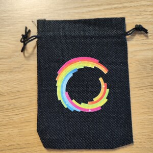 Puede incluir: Una peque&ntilde;a bolsa de cord&oacute;n negro con un dise&ntilde;o circular de arco&iacute;ris. El c&iacute;rculo est&aacute; formado por cuadrados de diferentes colores y tiene una estrella blanca en el centro.