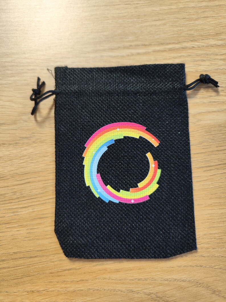 Puede incluir: Una peque&ntilde;a bolsa de cord&oacute;n negro con un dise&ntilde;o circular de arco&iacute;ris. El c&iacute;rculo est&aacute; formado por cuadrados de diferentes colores y tiene una estrella blanca en el centro.