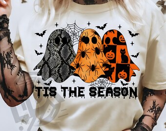 May include: Cream-colored t-shirt featuring three ghost designs with Halloween-themed patterns. The ghosts include a snakeskin print, orange with spiders, and a patchwork design with pumpkins and a black cat. The text "TIS THE SEASON" is printed below.